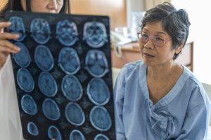 Older Asian woman with graying hair and in a light blue robe stares at numerous imaging scans of a brain that someone else in the background holds up for her