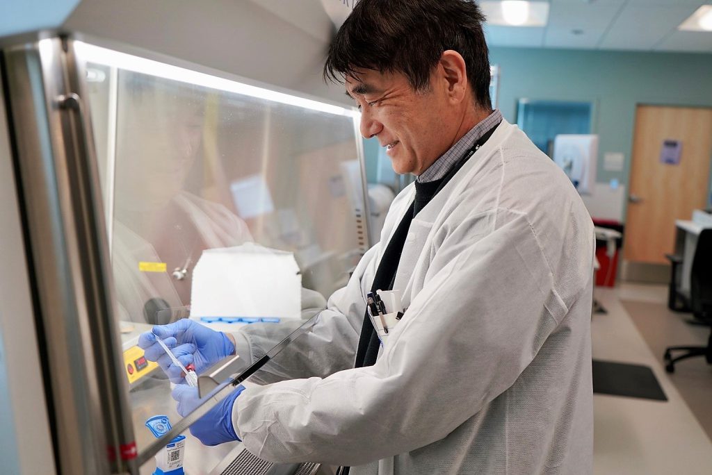 An Asian male lab scientist handles lab pipettes with samples outside of a safety shield piece of equipment in a hospital laboratory