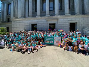 More than 100 people in teal T-shirts and rainbow accessories gather in front of gray building bricks and columns