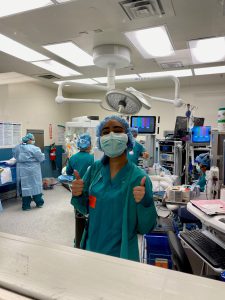 A female high school student in surgical scrubs and mask gets ready to observe a surgery in an operating room with other medical personnel.