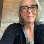 Caucasian woman with white bobbed hair, glasses, gray patterned blouse, silver necklace and black cardigan