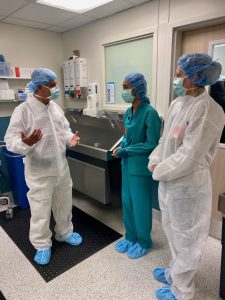 Three individuals wearing surgical scrubs and masks have a discussion near a hospital operating room