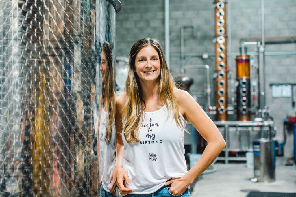 A Caucasian woman with long blonde hair in a white tank top and jeans stands in a gin distillery.
