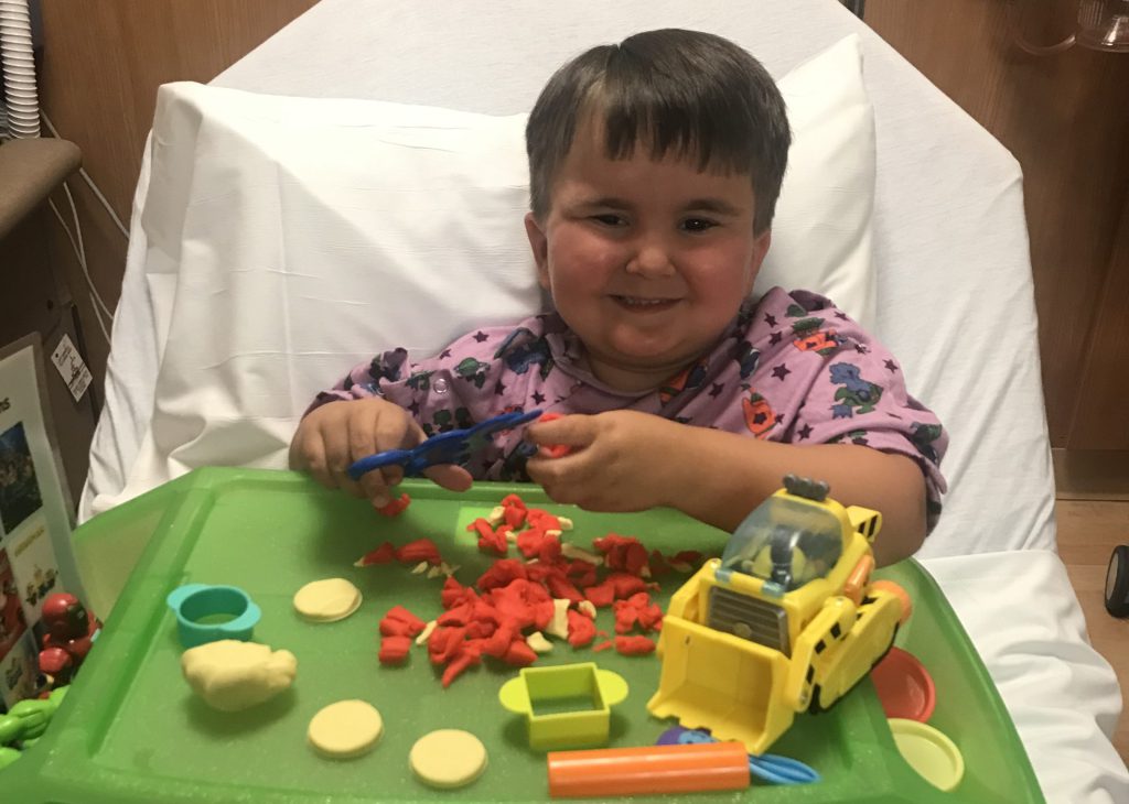A young Caucasian boy sits in a hospital bed playing with Play-Doh and various Play-Doh molds