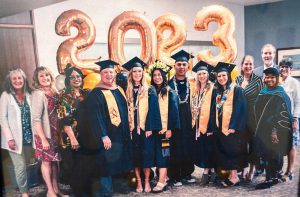 Group of people, including 6 who are wearing cap and gown to signify graduation, in front of balloons that represent the year 2023