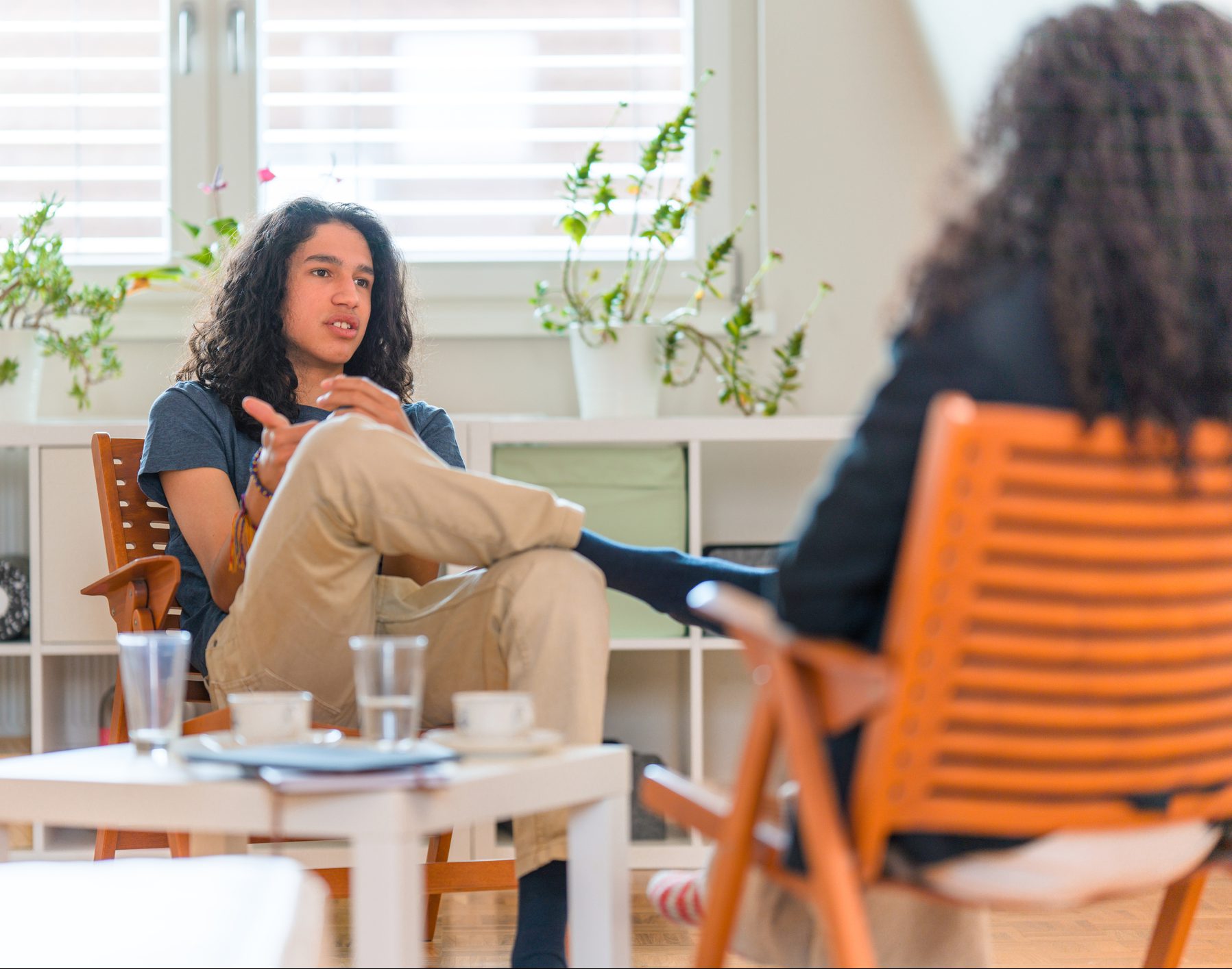 Young Hispanic man listening to mid adult woman mental health professional while sitting on sofa. They are conversing about his mental wellbeing.
