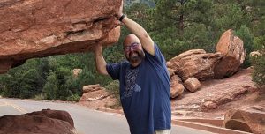 Caucasian male in blue T-shirt and glasses places hands on rock outcrop during hike
