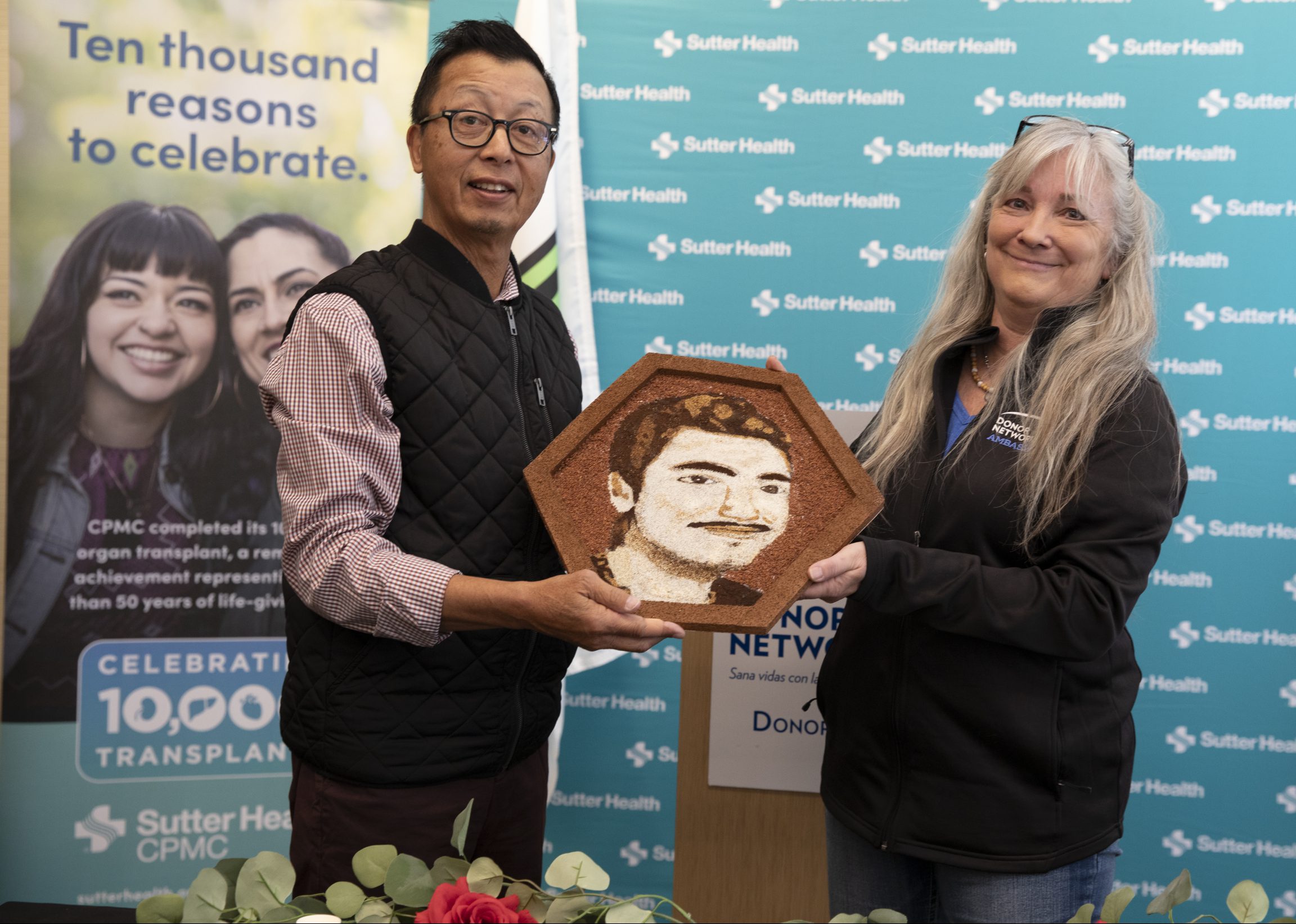 An Asian man and Caucasian woman hold a floragraph portrait of the woman's organ donor son.