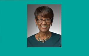 Black woman with short black hair, glasses, black/onyx jewelry and dark teal blouse