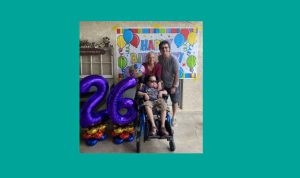 Mother, father and son who is in a wheelchair and sunglasses pose with a Happy Birthday banner and multicolored mylar balloons spelling out 26