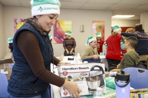A Latina woman gift wraps a handmixer wearing a teal Santa hat with Sutter Health logo while her colleagues socialize in the background.