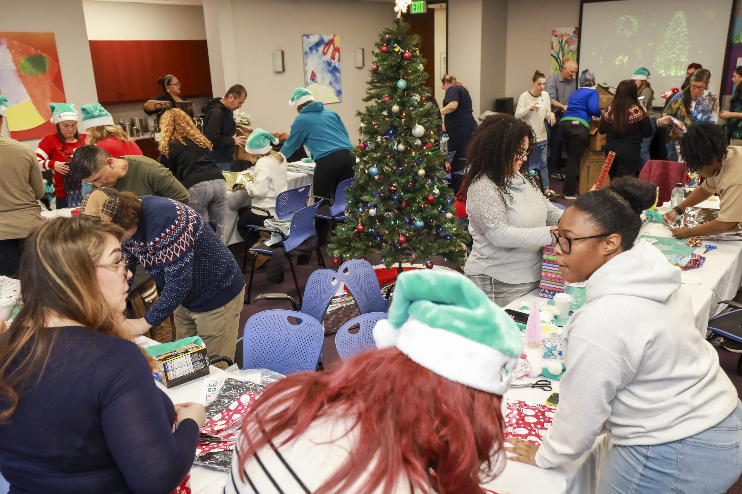 A large diverse group of adults wrap gifts in a conference room decorated for the winter holidays with a Christmas tree in the center of the room