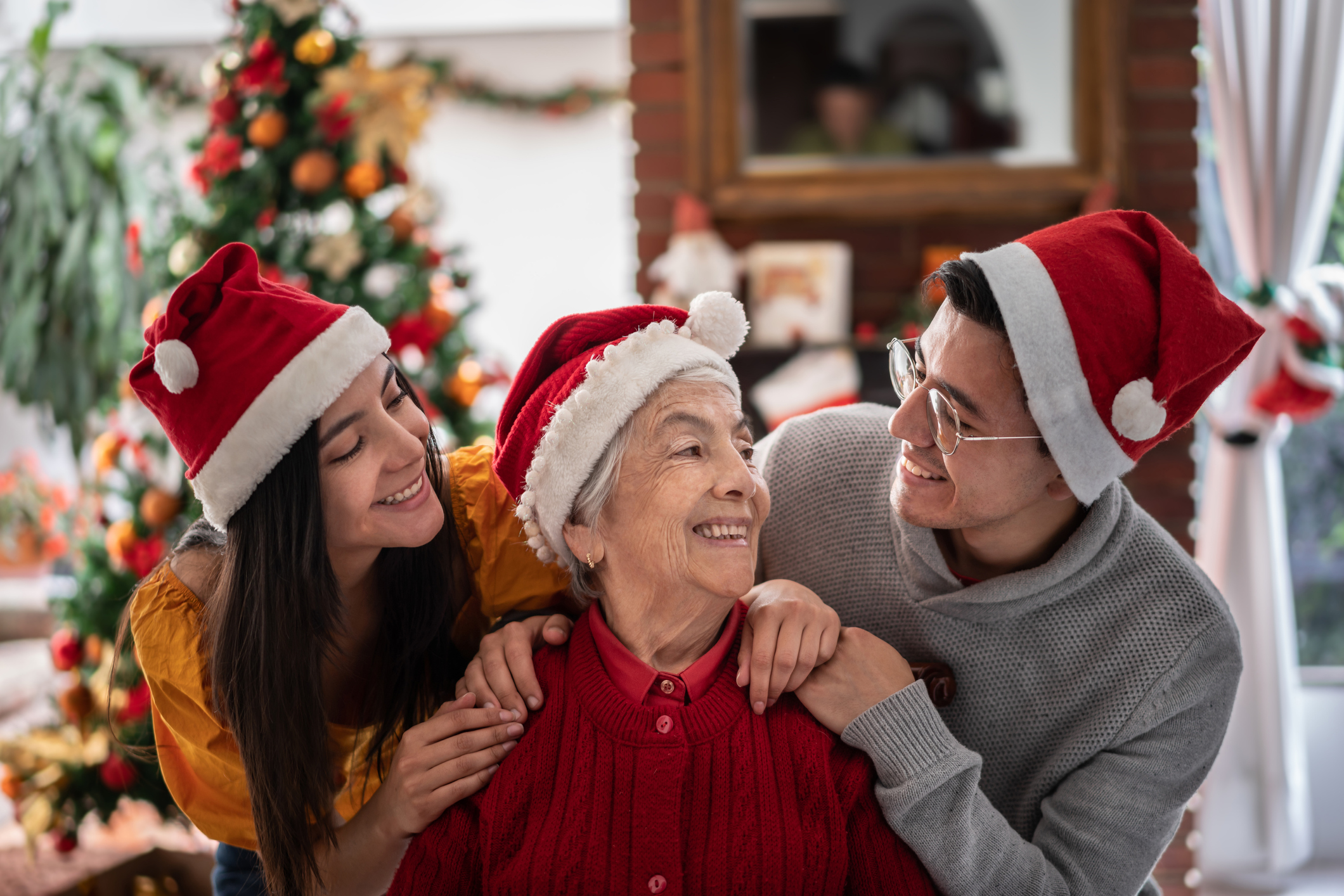Portrait of brother and sister hugging their grandmother looking at each other smiling all wearing Santa's hats on Christmas Eve - Celebration concepts