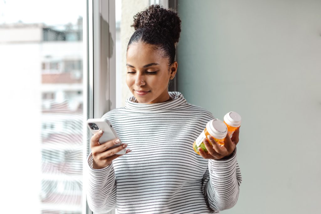 Black woman in striped turtleneck looks at her smartphone while holding two prescription bottles