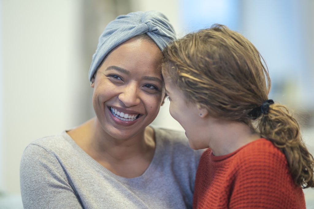 A black woman with cancer is spending time with her daughter. The woman is wearing a headscarf to hide her hair loss from chemotherapy treatment. The mother is holding her daughter. The two individuals are smiling and leaning in close with their foreheads touching.