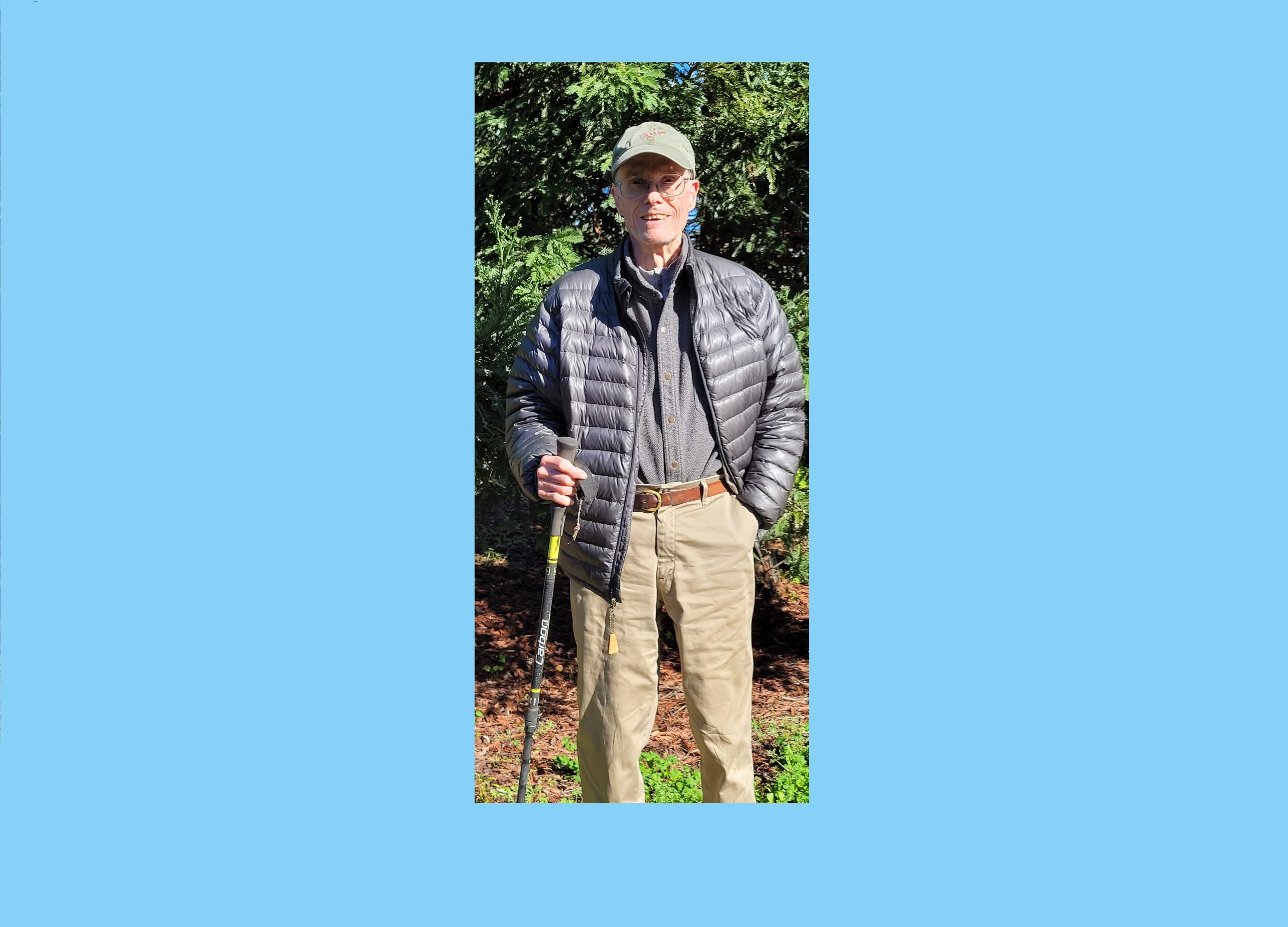 Older Caucasian man with tan baseball cap, charcoal gray puffy vest, charcoal gray long-sleeved shirt, khakis carrying a walking stick in front of evergreen trees with a blue border around the photo
