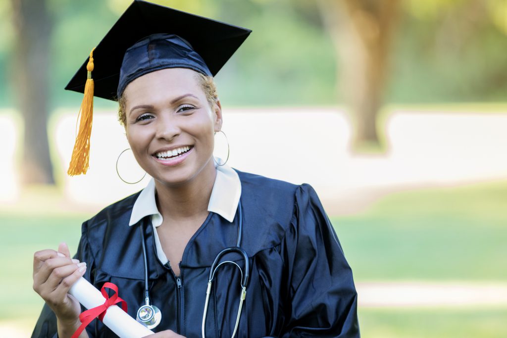 A Black female medical school graduate stands outdoors on a sunny day in her cap and gown, holding her rolled degree, and smiles for the camera. She also wears a stethoscope.