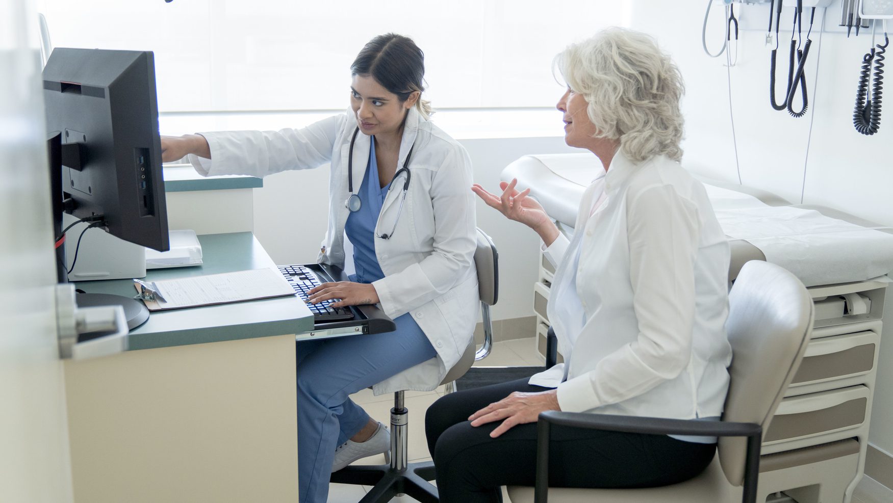 A Southeastern Asian female doctor sits inside an exam room and points to her computer while older Caucasian female sits next to her asking questions