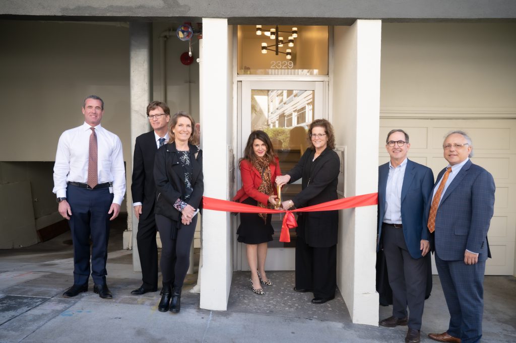 A group of individuals in business attire cut a red ribbon on a newly upgrade apartment building