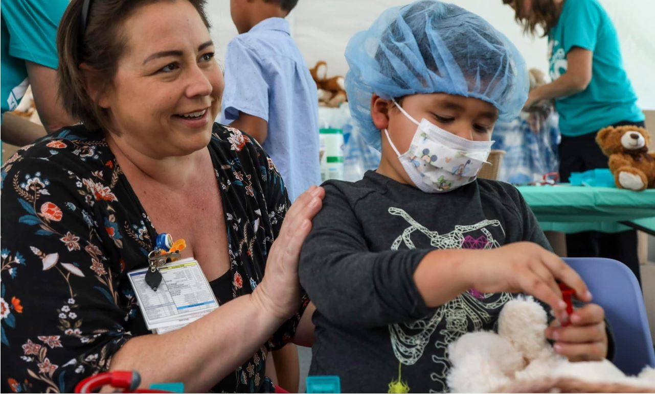 Latina clinician comforts young Latino boy with surgical mask and cap as he plays with a teddy bear