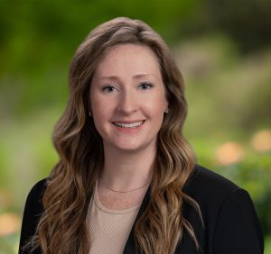 Caucasian woman with long strawberry blonde hair with a tan blouse and black blazer stands for a portrait in front of a greenery background