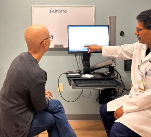 Bald woman with glasses speaks with male doctor with curly salt-and-pepper hair look at computer screen together while in an exam room.