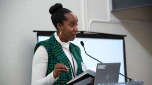A Black woman with a white turtleneck and green-checkered vest speaks at a podium