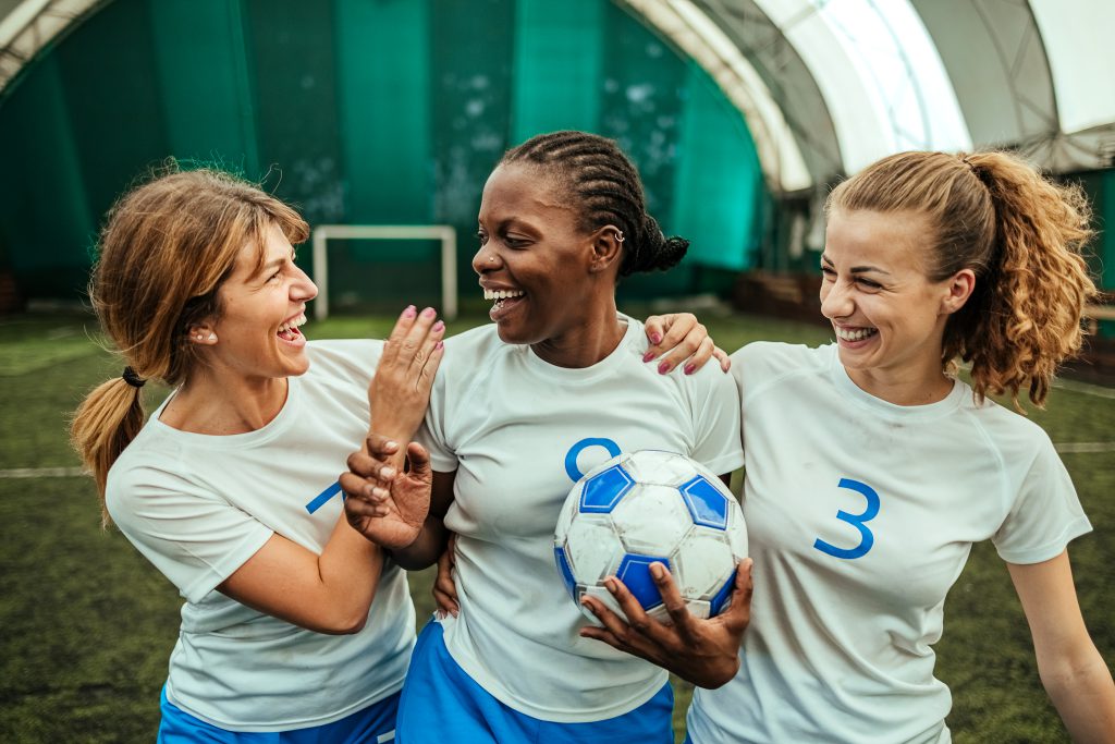 Three young women in white jerseys with blue numbers celebrate a soccer goal