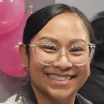 Asian woman with glasses and dark hair pulled into a bun smiles for the camera with pink balloons in the background