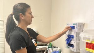 Caucasian woman with long, dark ponytail and dark-colored scrubs restocks plastic gloves inside clinic room