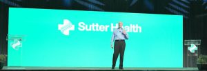 Bald, Caucasian man presenting to crowd with Sutter Health logo in background.