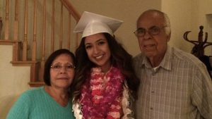 Latina woman in white graduation cap and gown poses with grandparents for photo in front of staircase