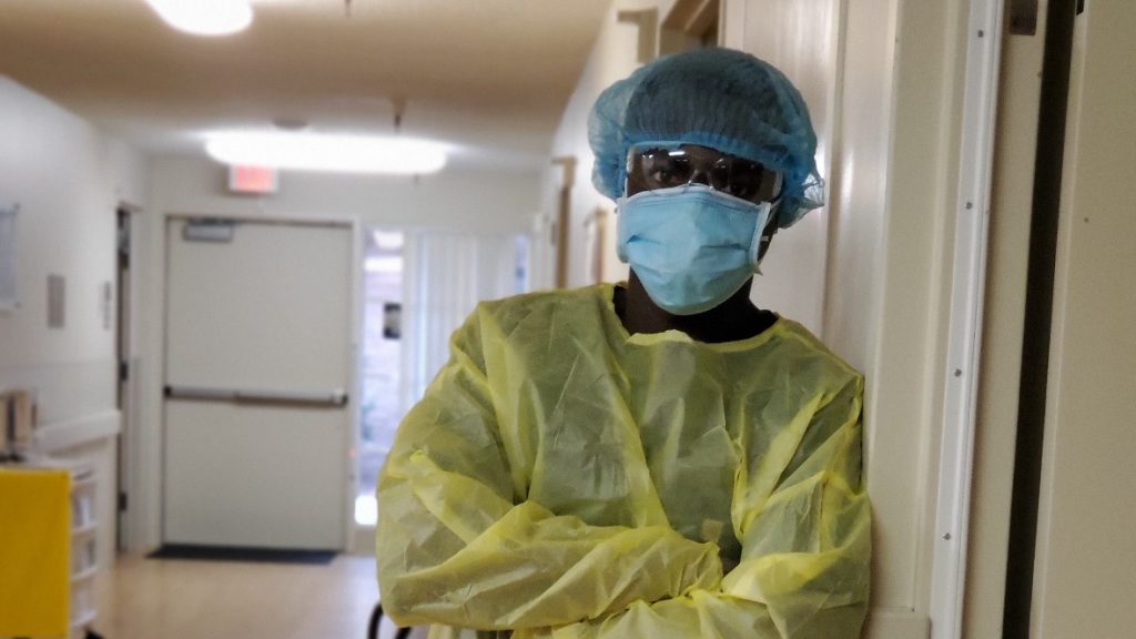 Black man in personal protective equipment including hair net, goggles, surgical mask and shield stands in the hallway of a hospital