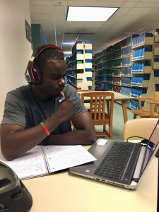 Black man with headphones stares at a laptop computer with notepad in front of him at a desk inside a library