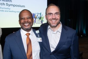 Two men smile for the camera while in front of a presentation screen.