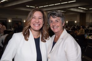 Two smiling women in white jackets posing for a photo in a conference setting.