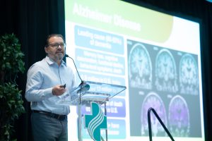 Caucasian man with graying beard and glasses stands at clear podium with large screen behind him projecting brain scans