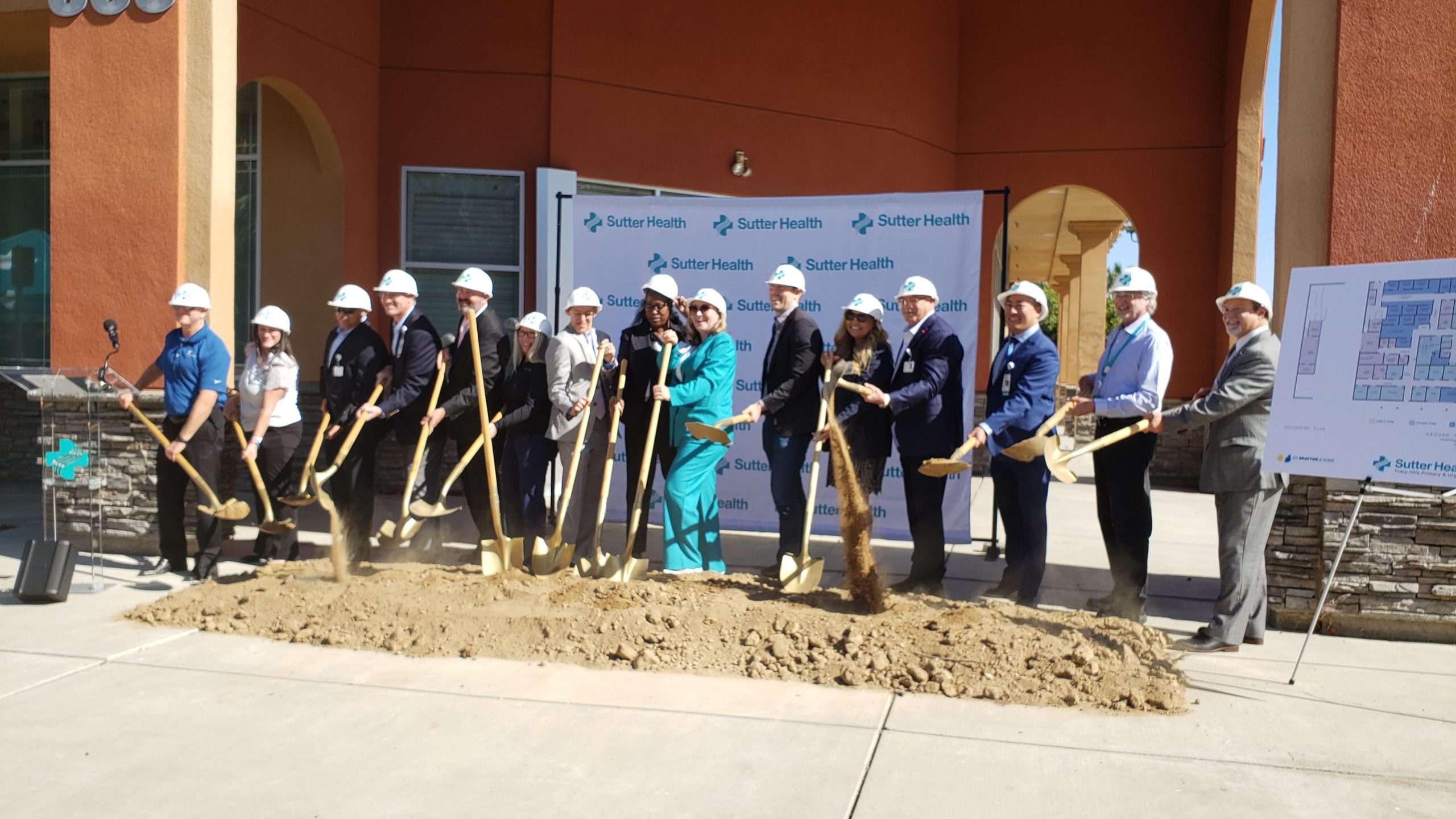 Numerous adults with hard hats and shovels stand behind pile of dirt with some shoveling