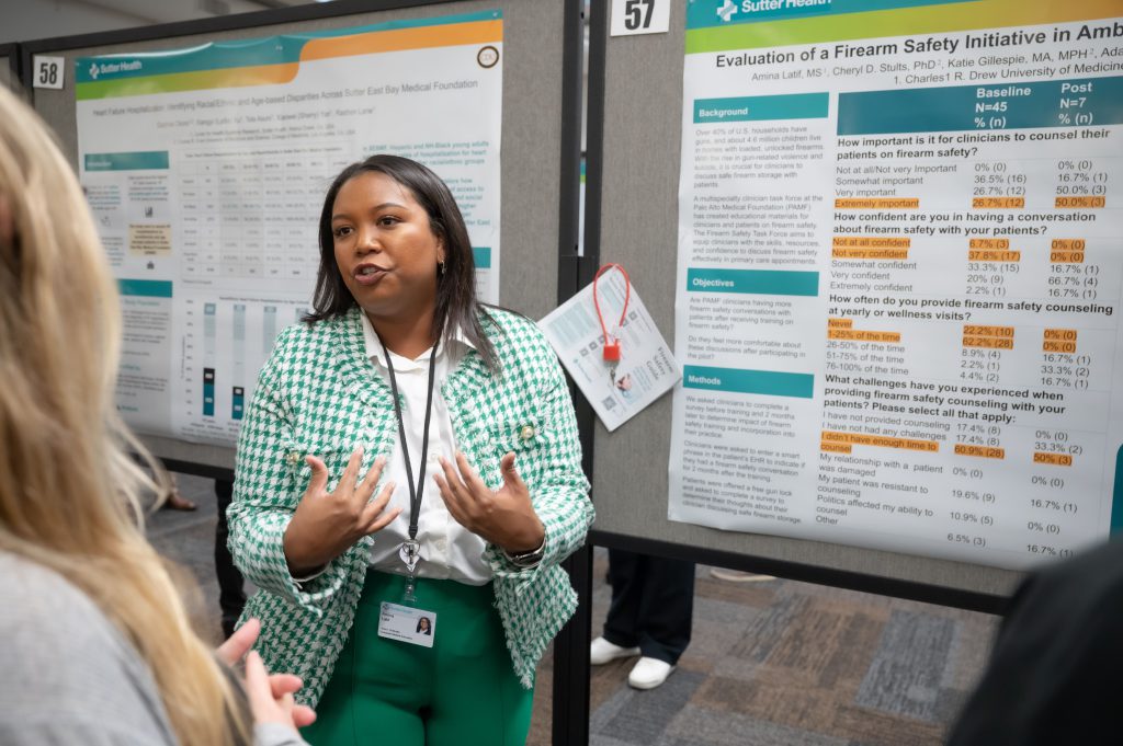 Black woman with green and white houndstooth-patterned jacket with pearl embellishments and green pants stands in front of poster speaking to blond haired woman with her back to the camera