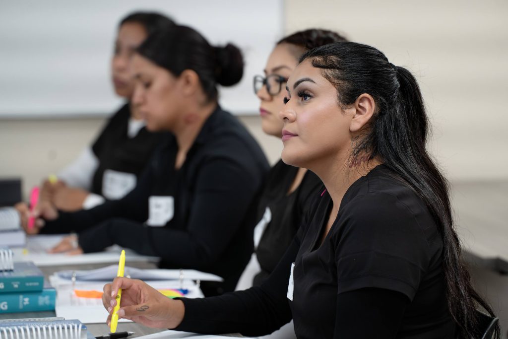 Four Latina women in a classroom sitting at a long desk and wearing black scrubs with a dry erase board in the background