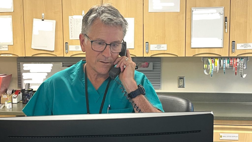 Man with salt and pepper hair and glasses talks on the phone in front of a computer monitor with rows of cabinets behind him