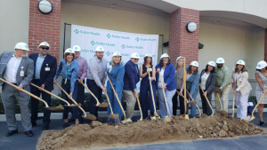 Sutter Health executives, city, county, and national government representatives at the ceremonial groundbreaking. Each person in a line with a shovel in front of dirt posing for a photo.