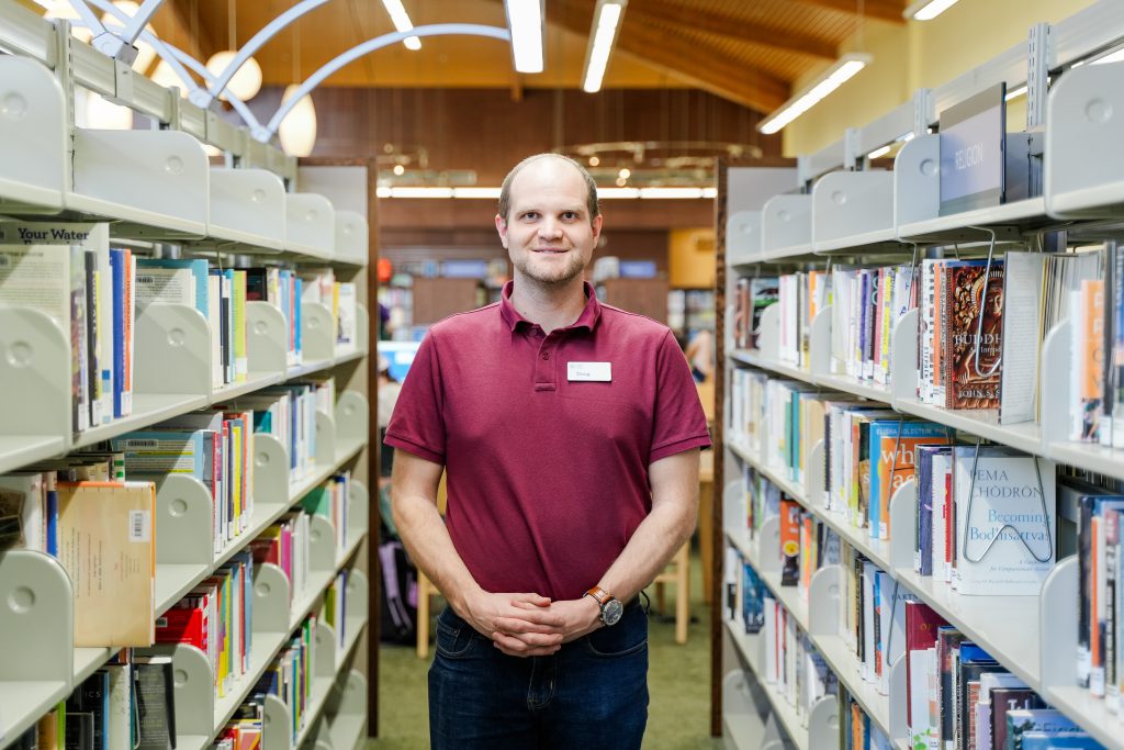 Douglas Sheehan, a white male, is wearing a maroon shirt and dark pants. He is standing in the middle of an aisle, surrounded by rows of files and books.