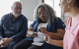 An African American woman is sitting down. She is wearing a white shirt and dark pants. She has grey hair and glasses. She is pricking her finger with an older white male sitting next to her. He also has glasses.