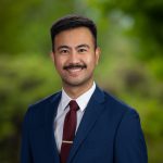 Asian man with dark hair and mustache and blue suit jacket white shirt and red tie poses in front of greenery background