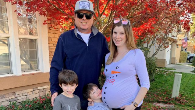 Man, pregnant woman and two toddler boys smile for posed photo in their front yard with orange and red foliage surrounding them