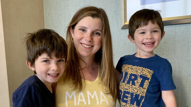 Two young boys with brown shaggy hair and navy t-shirts huddle on either side of their mother with long brownish blonde hair who is pregnant with a yellow t-shirt on