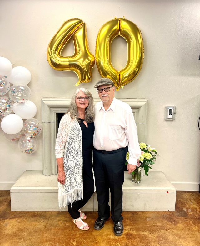 Older white couple stand in front of their fireplace and mantle with gold, white and silver balloons behind them.