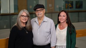 A trio of adults pose for with a balcony and glass walls behind them and artwork on walls in the distance