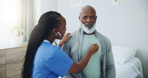 Older Black Male and nurse with listening on stethoscope for heartbeat assessment or cardiovascular health on bed. Elderly patient, doctor and cardiology in hospital for lung disease or chest infection